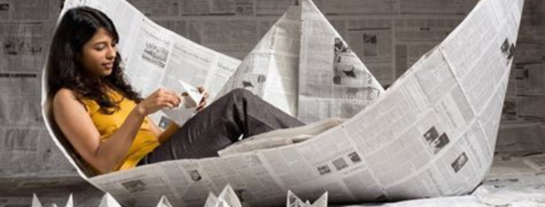 Una giovane donna siede rilassata all'interno di una grande barca di carta fatta di giornali e piega una barchetta, evocando il relax e il distacco dallo stress quotidiano